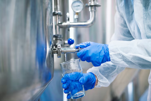 Close up view of hands in protective blue colour gloves opening a tap valve and filling the glass with samples from industrial machine reservoir